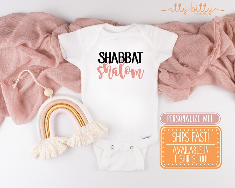 May include: White baby onesie with the text "Shabbat Shalom" in black and pink lettering. The onesie is on a white background with a pink fabric and a rainbow-coloured yarn wall hanging.