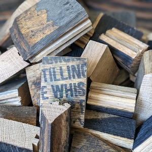 May include: A close-up of a pile of reclaimed wood pieces, some of which are stained with a dark brown color. The wood pieces are arranged in a random pattern, and some of them have the words "Tilling & Whis" printed on them.