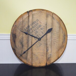 May include: A wooden clock with black hands, made from a repurposed whiskey barrel. The barrel has the text "J.B. Beam Brands Co. Bourbon Whiskey 100 Proof R.C. 53 Gal. C.Y. 230" printed on it.
