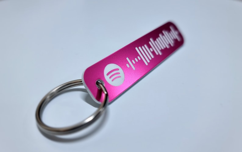Pode incluir: Um chaveiro de metal rosa com um logotipo Spotify branco e um c&oacute;digo de barras branco. O chaveiro est&aacute; preso a um anel de metal prateado.