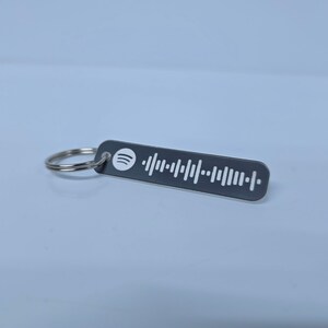 Pode incluir: Um chaveiro de metal preto com um logotipo Spotify branco e um c&oacute;digo de barras branco. O chaveiro est&aacute; preso a um anel prateado.