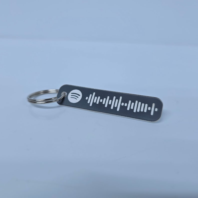Pode incluir: Um chaveiro de metal preto com um logotipo Spotify branco e um c&oacute;digo de barras branco. O chaveiro est&aacute; preso a um anel prateado.