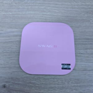 Pu&ograve; includere: Un oggetto quadrato lilla con angoli arrotondati. Il testo "SWAG II" &egrave; stampato al centro. Un'etichetta "Parental Advisory Explicit Content" si trova nell'angolo in basso a destra. L'oggetto &egrave; su una superficie di legno grigio.