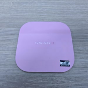 Pu&ograve; includere: Un sottobicchiere quadrato lilla con angoli arrotondati. Il testo "SWAG II" &egrave; stampato al centro. Un'etichetta in bianco e nero "Parental Advisory Explicit Content" si trova nell'angolo in basso a destra.