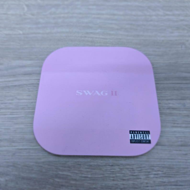 Pu&ograve; includere: Un sottobicchiere quadrato lilla con angoli arrotondati. Il testo "SWAG II" &egrave; stampato al centro. Un'etichetta in bianco e nero "Parental Advisory Explicit Content" si trova nell'angolo in basso a destra.