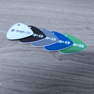 May include: A selection of six guitar plectrums in white, black, grey, blue, and green. Each plectrum has a white soundwave design. The plectrums are displayed on a wooden surface.