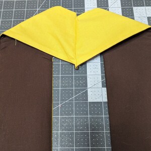 Puede incluir: Un tri&aacute;ngulo de tela amarilla cosido a dos tiras de tela marr&oacute;n. La tela est&aacute; colocada sobre una estera de corte gris.