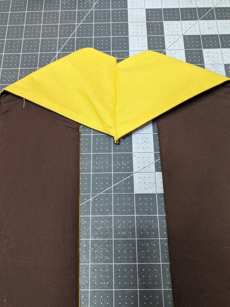 Puede incluir: Un tri&aacute;ngulo de tela amarilla cosido a dos tiras de tela marr&oacute;n. La tela est&aacute; colocada sobre una estera de corte gris.