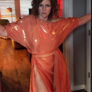 May include: A woman wearing a shiny, peach-colored, sleeveless dress with a sash tied at the waist. The dress has a high slit on the side.