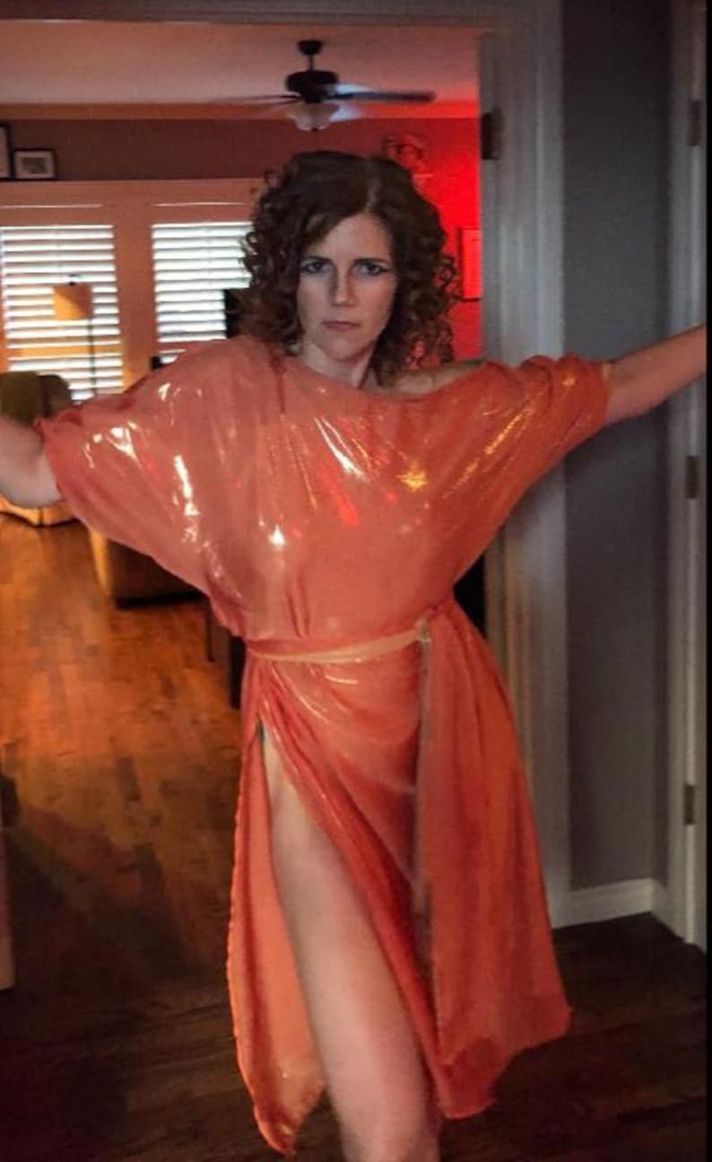 May include: A woman wearing a shiny, peach-colored, sleeveless dress with a sash tied at the waist. The dress has a high slit on the side.