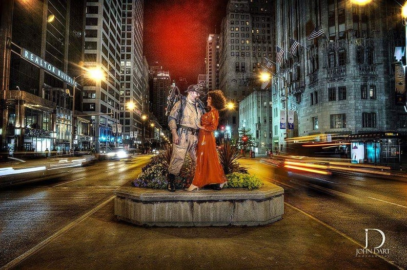 May include: A couple dressed in a vintage style pose in front of a city skyline at night. The man is wearing a brown leather jacket and the woman is wearing a red dress. The couple is standing in front of a planter with green plants.