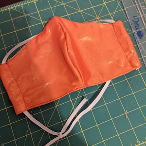 May include: A bright orange face mask with white elastic straps. The mask is made of a shiny fabric and has a pleated design.