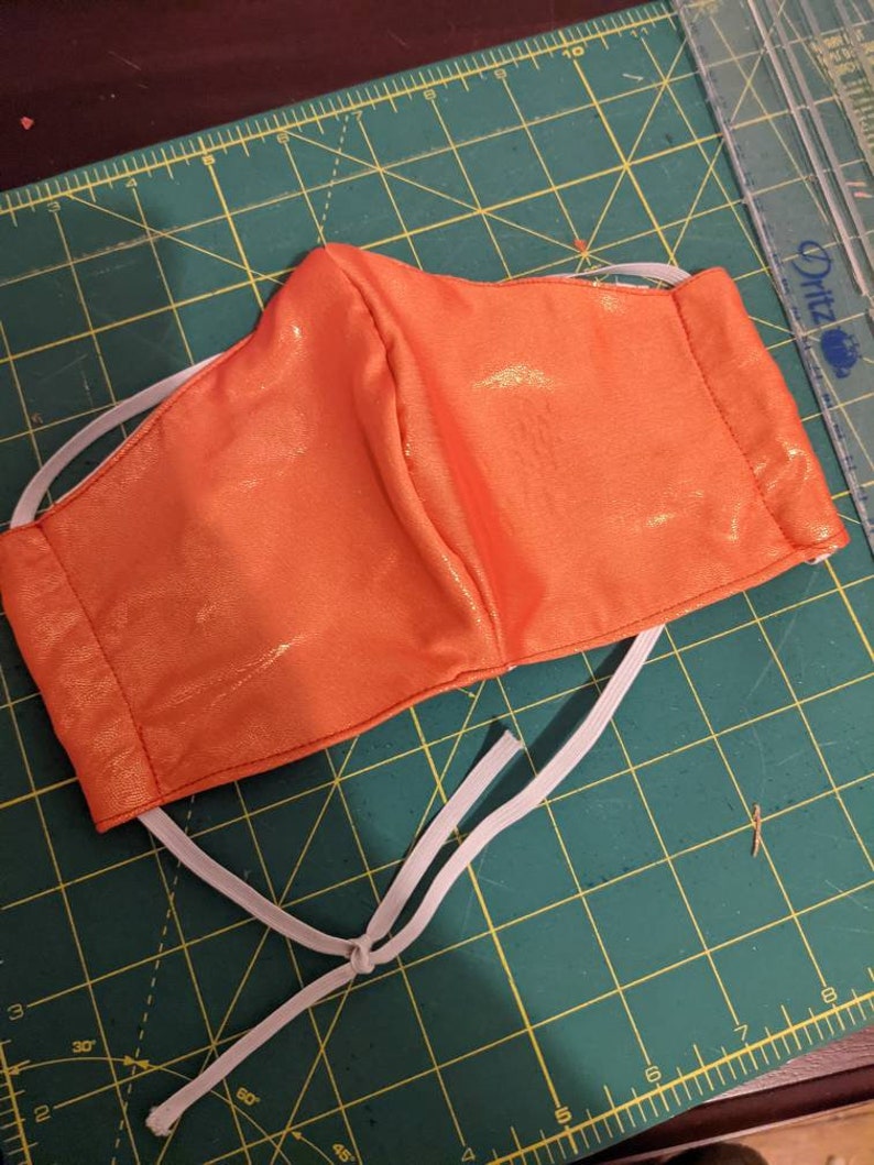May include: A bright orange face mask with white elastic straps. The mask is made of a shiny fabric and has a pleated design.