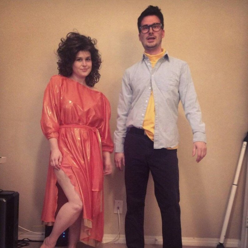 May include: A woman in a bright orange dress with a yellow belt stands next to a man wearing a light blue button-down shirt over a yellow shirt and dark pants.