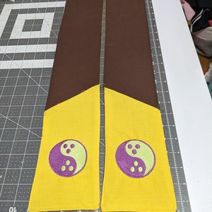 Puede incluir: Dos tiras de tela marr&oacute;n con extremos amarillos y s&iacute;mbolos yin yang morados y verdes bordados en ellas.