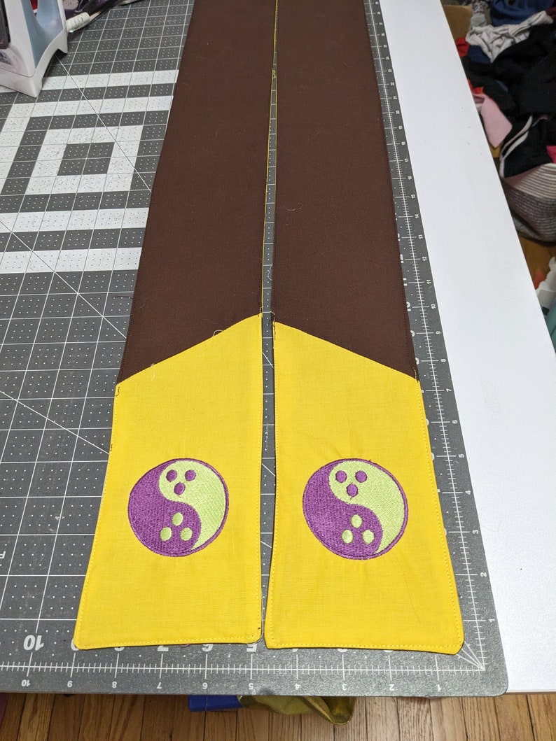 Puede incluir: Dos tiras de tela marr&oacute;n con extremos amarillos y s&iacute;mbolos yin yang morados y verdes bordados en ellas.