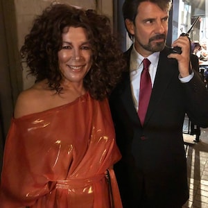 May include: A woman in a bright orange dress with a man in a black suit and a red tie. The man is holding a walkie-talkie.