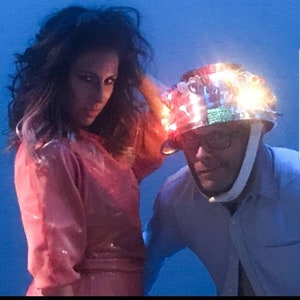 May include: A woman in a pink outfit stands next to a man wearing a silver helmet decorated with colorful lights. The helmet has a white strap and the man is wearing glasses.