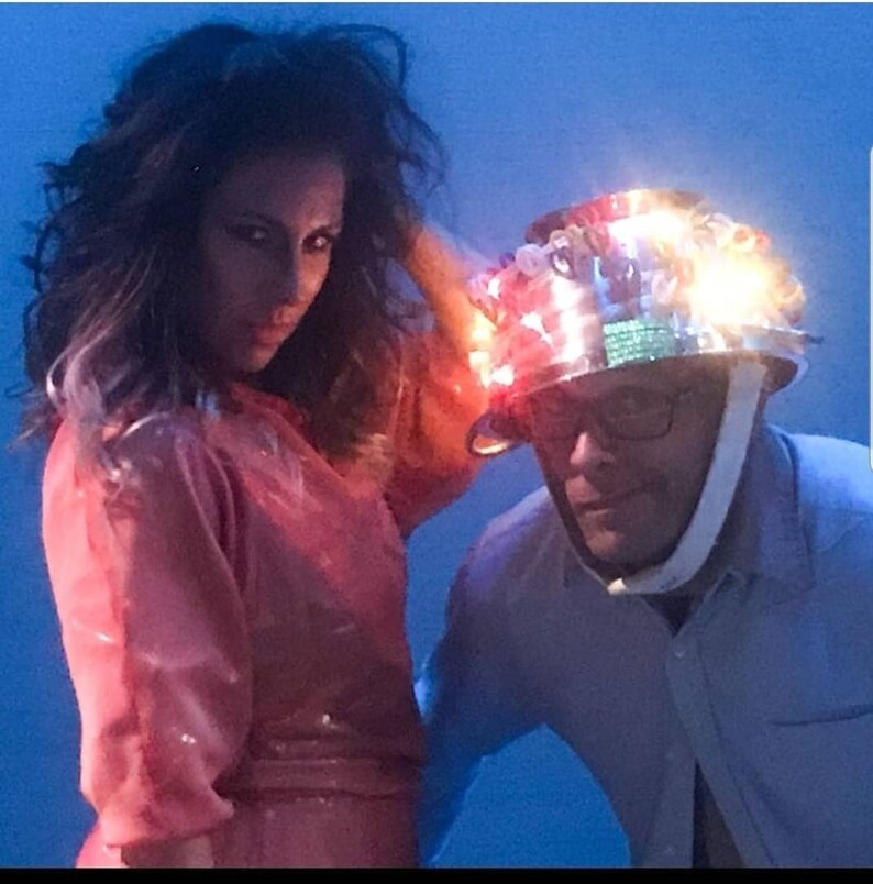 May include: A woman in a pink outfit stands next to a man wearing a silver helmet decorated with colorful lights. The helmet has a white strap and the man is wearing glasses.