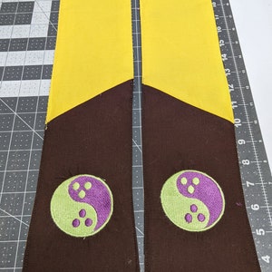 Puede incluir: Dos fajas de tela amarilla y marr&oacute;n con un s&iacute;mbolo yin yang verde y morado bordado en cada una.