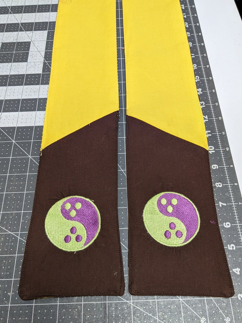 Puede incluir: Dos fajas de tela amarilla y marr&oacute;n con un s&iacute;mbolo yin yang verde y morado bordado en cada una.