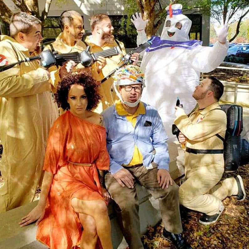 May include: A group of people dressed in costumes, including a Ghostbusters costume, a Stay Puft Marshmallow Man costume, and a woman in a bright orange dress. The group is posing for a photo, with the Stay Puft Marshmallow Man in the center.