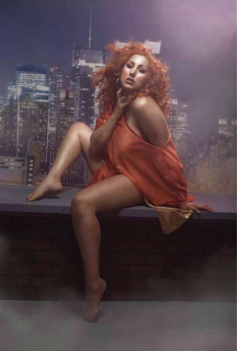 May include: A woman with red curly hair is sitting on a ledge with a city skyline in the background. She is wearing a loose-fitting orange top and has one shoulder exposed.
