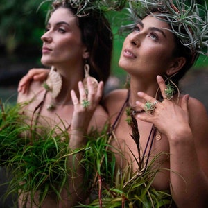 May include: Two people adorned with plant-based accessories, including headpieces and jewelry. They wear green plant arrangements as tops. The setting is outdoors, with a blurred background of greenery and water, creating a natural, artistic aesthetic.