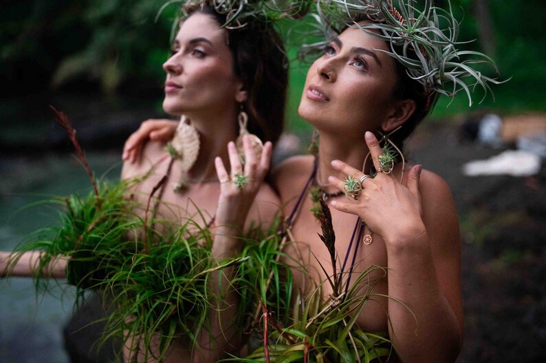 May include: Two people adorned with plant-based accessories, including headpieces and jewelry. They wear green plant arrangements as tops. The setting is outdoors, with a blurred background of greenery and water, creating a natural, artistic aesthetic.