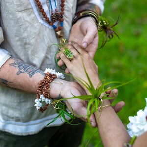 May include: Close-up of two people holding hands, adorned with natural jewelry. One person wears a ring with a small plant, and both have air plants attached to their wrists. The scene suggests a bohemian or nature-inspired theme.
