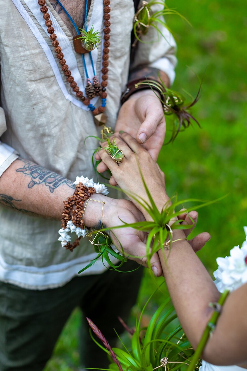 May include: Close-up of two people holding hands, adorned with natural jewelry. One person wears a ring with a small plant, and both have air plants attached to their wrists. The scene suggests a bohemian or nature-inspired theme.