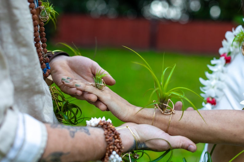 May include: A close-up of two hands, one wearing a ring and the other a bracelet, each holding a small air plant in a gold wire frame. The background is a blurred green lawn.