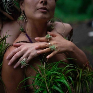 May include: A close-up shot of two people adorned with jewelry and plants. The hands of one person are adorned with rings featuring a plant design. The jewelry and plants create a natural, earthy aesthetic.