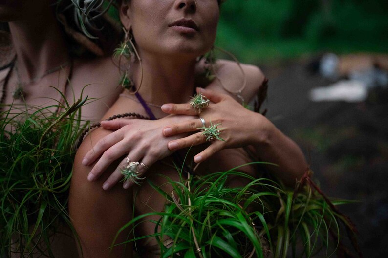 May include: A close-up shot of two people adorned with jewelry and plants. The hands of one person are adorned with rings featuring a plant design. The jewelry and plants create a natural, earthy aesthetic.