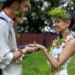 May include: A couple exchanging rings during an outdoor ceremony. The woman wears a white dress and a floral garland. The man wears a light-colored shirt and a hat. Both have air plants as accessories, creating a natural, bohemian aesthetic.