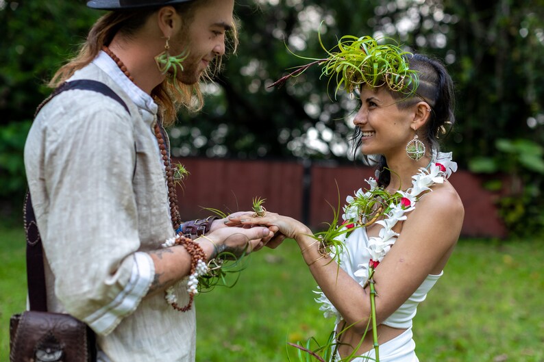 May include: A couple exchanging rings during an outdoor ceremony. The woman wears a white dress and a floral garland. The man wears a light-colored shirt and a hat. Both have air plants as accessories, creating a natural, bohemian aesthetic.