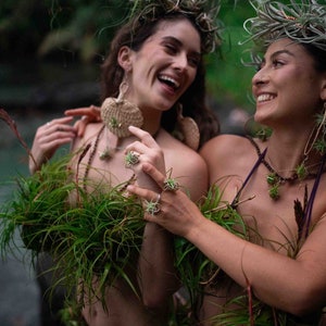 May include: Two people wearing plant-based jewelry and accessories, including earrings, rings, and necklaces. They are adorned with air plants and greenery, with crowns of plants in their hair. The setting appears to be outdoors, with a natural, green backdrop.