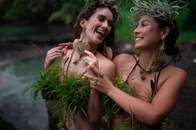 May include: Two people wearing plant-based jewelry and accessories, including earrings, rings, and necklaces. They are adorned with air plants and greenery, with crowns of plants in their hair. The setting appears to be outdoors, with a natural, green backdrop.