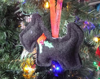 May include: A black felt Christmas ornament shaped like a dog with a red and green holly sprig. The ornament is hanging on a Christmas tree with red and green lights.