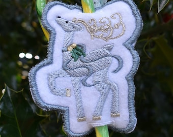Puede incluir: Adorno navideño con un ciervo de fieltro blanco con bordados plateados y detalles dorados. El adorno está sujeto a un bastón de caramelo verde y amarillo con una cinta azul claro. El fondo es un arbusto de acebo.