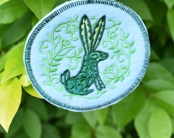 May include: A blue ribbon hangs from a green leafy branch, attached to a round blue felt ornament with a green embroidered rabbit design.