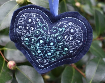 Puede incluir: Un adorno en forma de corazón de fieltro azul con un diseño bordado en blanco y azul. El adorno está colgado de una cinta azul.
