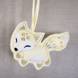 May include: A white felt fox ornament with gold and silver embroidery. The fox has a closed eye and a small smile. The ornament is hanging from a yellow ribbon.