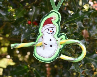 May include: A green felt snowman ornament with a red hat and a candy cane. The snowman is holding the candy cane in its arms.