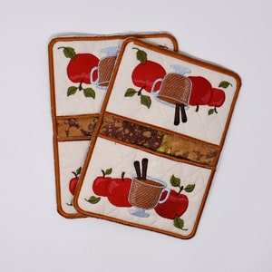 May include: Two white quilted pot holders with brown trim feature an embroidered design of apples and a mug of hot cider with cinnamon sticks.