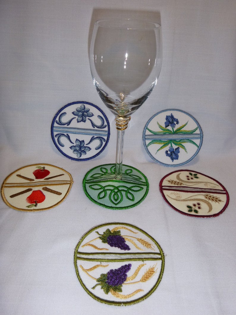May include: Six embroidered round coasters with various designs, including a red apple with cinnamon sticks, a green Celtic knot, a purple grape with wheat stalks, a blue flower, and a white flower. The coasters are arranged around a clear wine glass.
