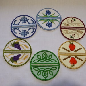 May include: Six round embroidered fabric coasters with different designs. The designs include a grapevine and wheat, an apple and cinnamon sticks, a green Celtic knot, a blue iris, a red berry and wheat, and a blue flower.