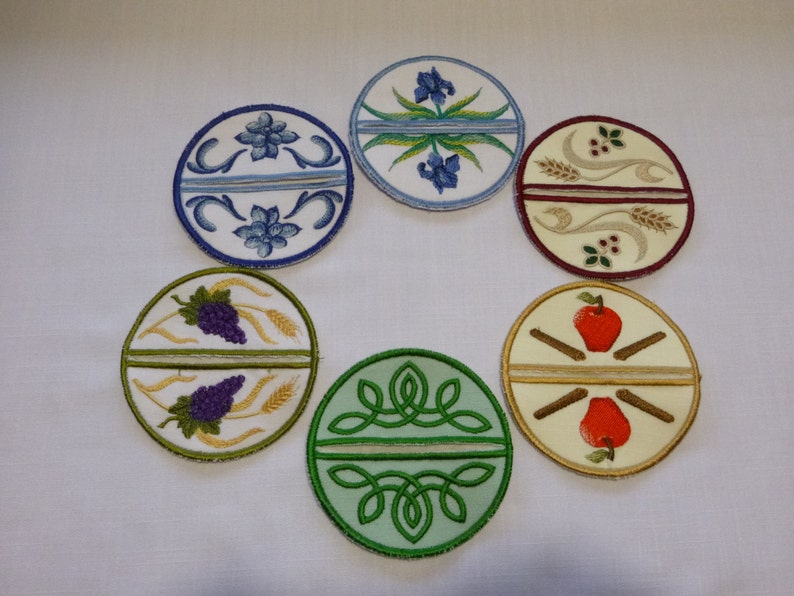 May include: Six round embroidered fabric coasters with different designs. The designs include a grapevine and wheat, an apple and cinnamon sticks, a green Celtic knot, a blue iris, a red berry and wheat, and a blue flower.