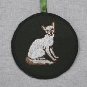 Op de afbeelding: Een ronde, donkergroene geborduurde patch met een witte kat met bruine accenten, zittend. De patch heeft een zwarte rand en een groen lint om op te hangen. De kat is gedetailleerd met wit garen.