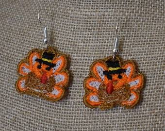 May include: A pair of dangle earrings featuring embroidered turkey designs. The turkeys are brown, orange, and white with black hats and red wattles. The earrings are silver.
